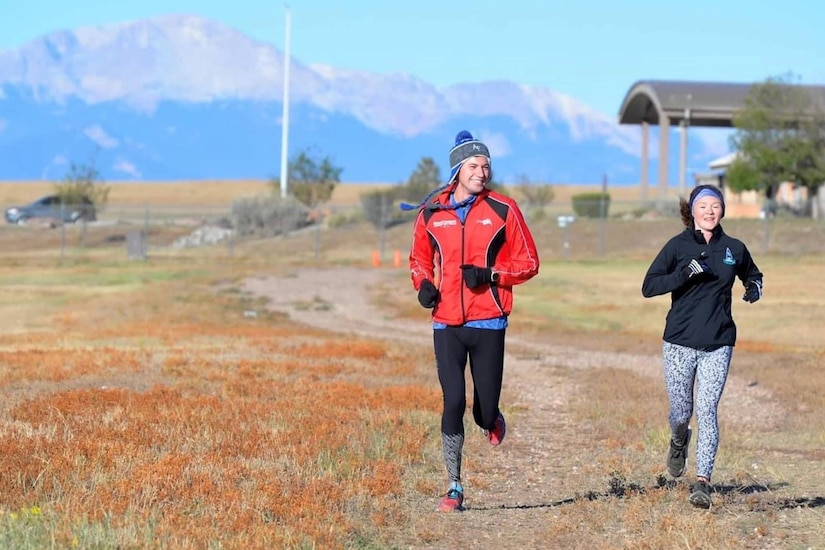 Two people run in a field. A mountain range can be seen behind them.