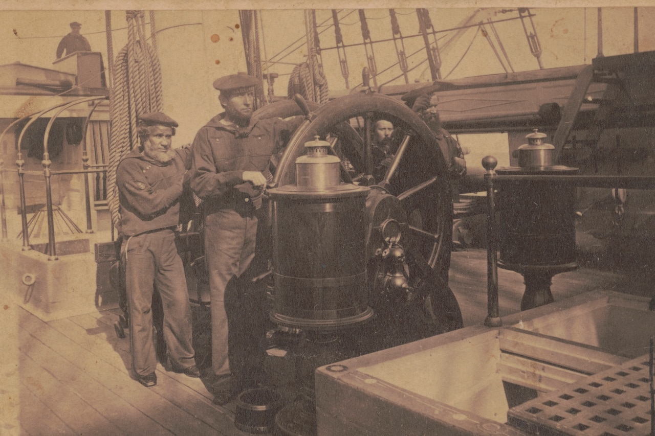 Two men stand behind the helm of a ship.