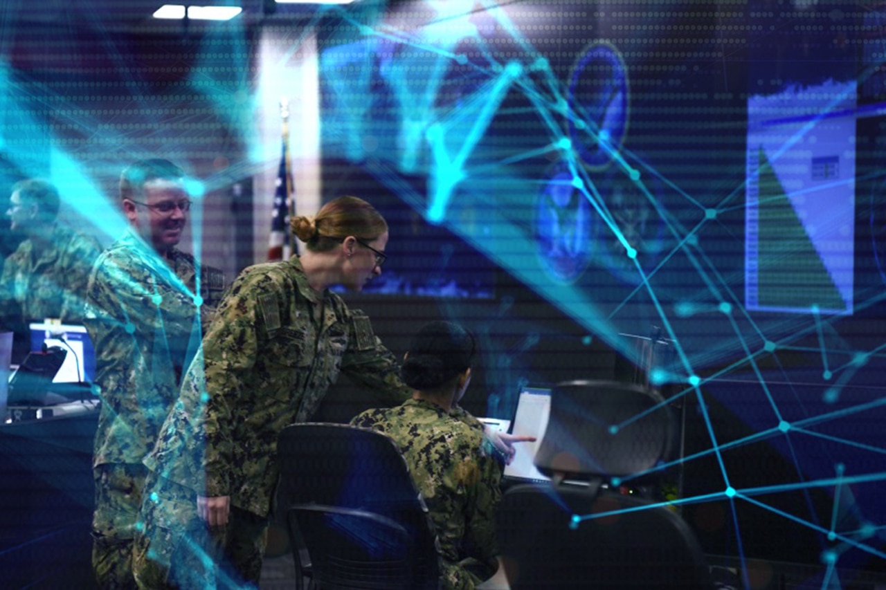 Uniformed personnel look at a computer screen. The wall has large video screens.