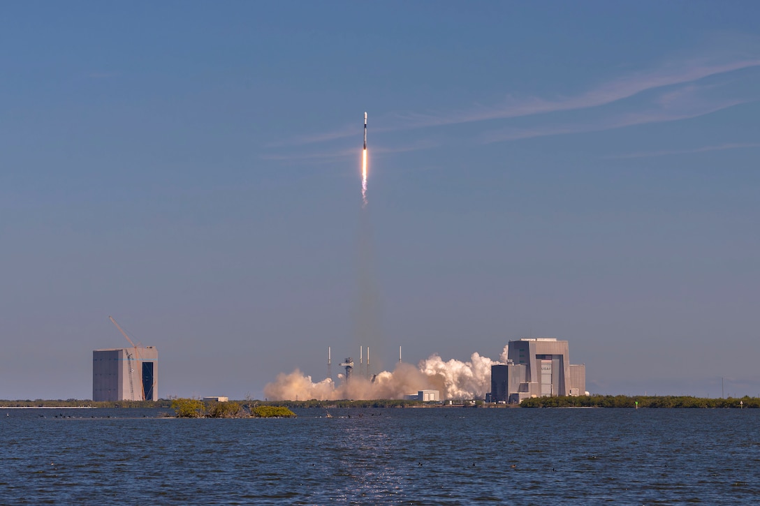 Smoke arises as a rocket launches into the sky with a body of water in the foreground.