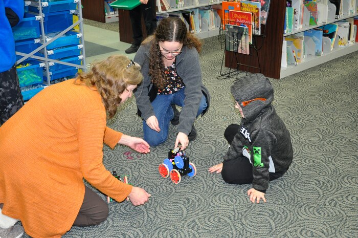 STEM outreach mentors from U.S. Navy Strategic Systems Programs (SSP) kneel on the floor to help a young student test his vehicle design during a Science, Technology, Engineering, and Math (STEM) outreach event at the Camden County Library. SSP Strategic Weapons Facility Atlantic (SWFLANT) STEM coordinator Caren Spahr (left) and SWFLANT Mentor Rebecca Pasquale (middle) both evaluated the student's design. SWFLANT donated a STEM cart originally given to Camden County High School containing all the educational materials for this event. Employees from SSP sites, like SWFLANT, are heavily involved in nationwide naval STEM outreach, volunteering to provide mentorship and sponsorship in local communities where their sites or commands are located.