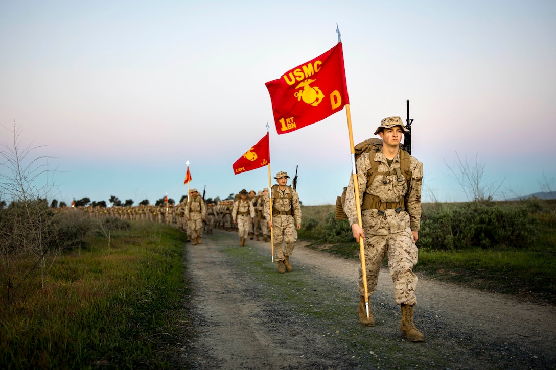 Two Marine Corps recruits holding red flags lead dozens of recruits walking in formation on a dirt road between a field under a pinkish sky.