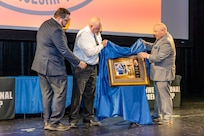 Kyle Weir and Reuben Farley unveil a plaque for apprentice Hall of Fame inductee Allan Schott, training instructor, Shop 38, Marine Machinist, Jan. 18, 2024, during a graduation ceremony at Southwestern College, in Chula Vista, Calif. (U.S. Navy photos by Jeb Fach)