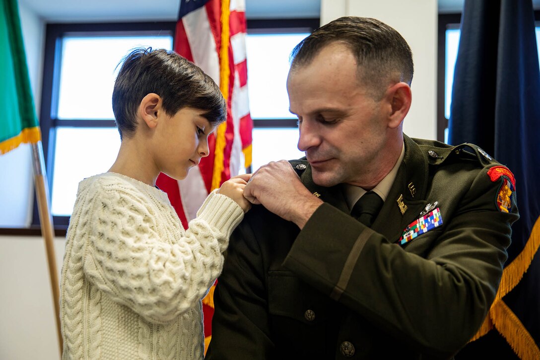 A child pins rank insignia on a shoulder of the uniform of his kneeling father.