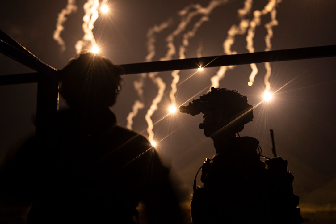 Two Marines are silhouetted against a dark sky as aerial range firing takes place in front of them.