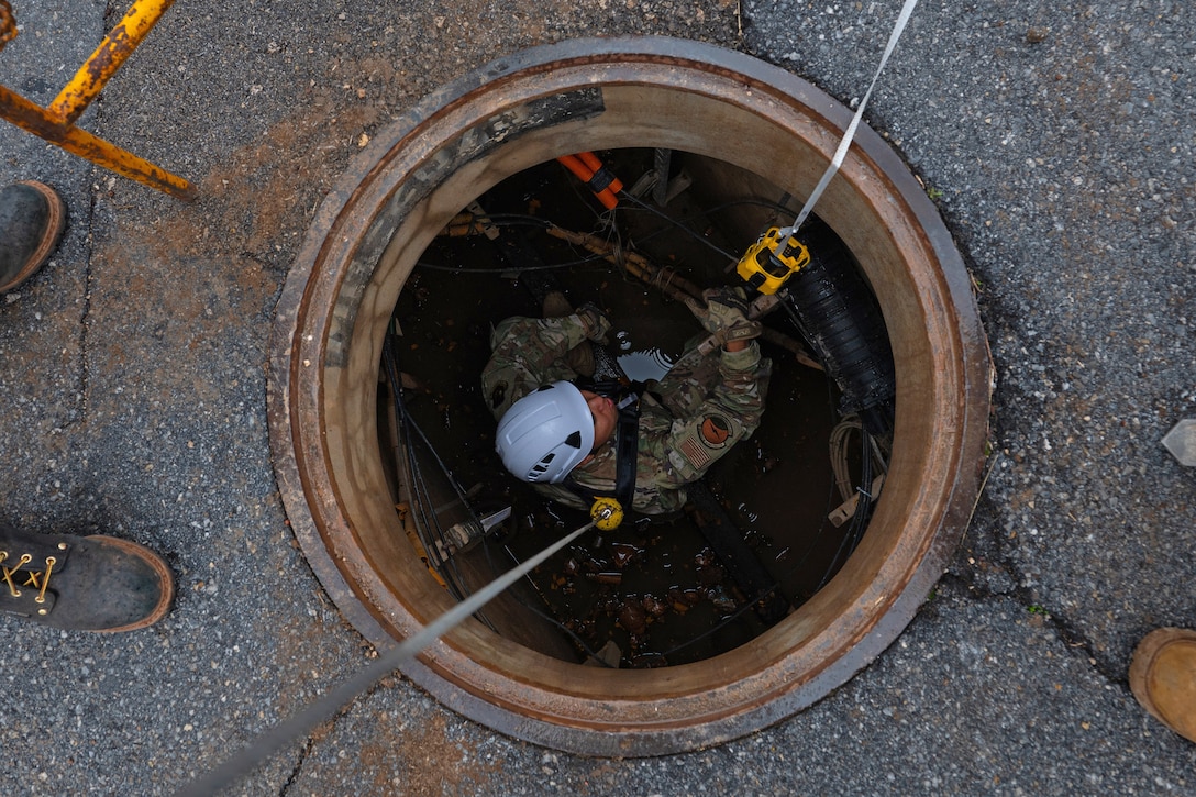 An airman wears a helmet inside of a manhole as the boots of two other people can be seen surrounding the manhole.