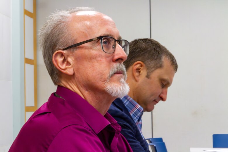 Man in a burgundy shirt and a grey gotee and glasses stands in front of another man in a blue plaid shirt.