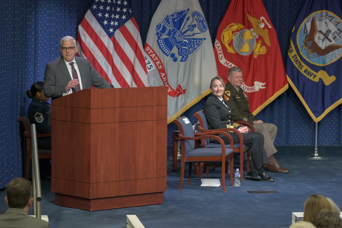 Assistant Secretary of Defense for Sustainment Christopher Lowman speaks from behind a lectern with VADM Michelle Skubic and LTG Mark Simerly looking on