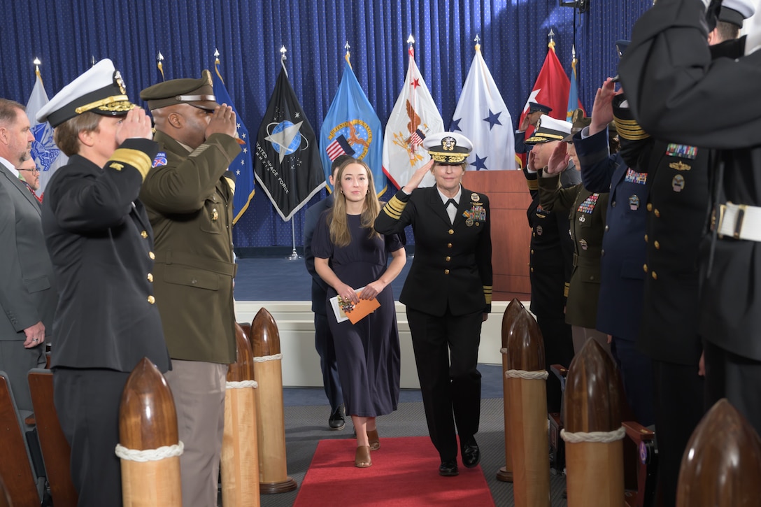 VADM Skubic and her family proceed down an aisle flanked by saluting sideboys with military service flags on a stage behind her