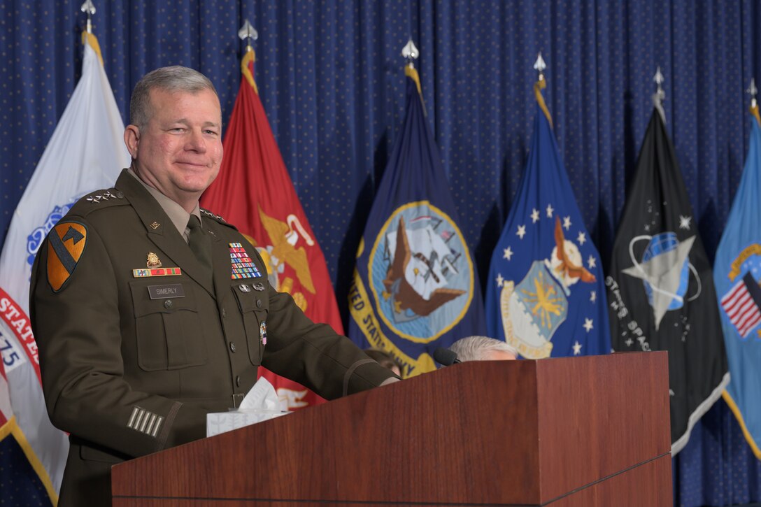 LTG Mark Simerly speaks from behind a lectern on a stage with military service flags in the background