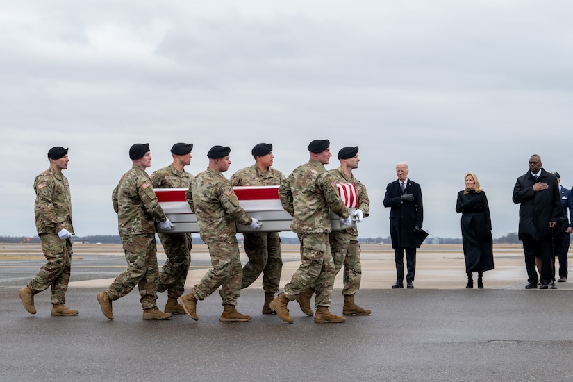 Three civilians watch as soldiers carry a flag-draped coffin from a plane.