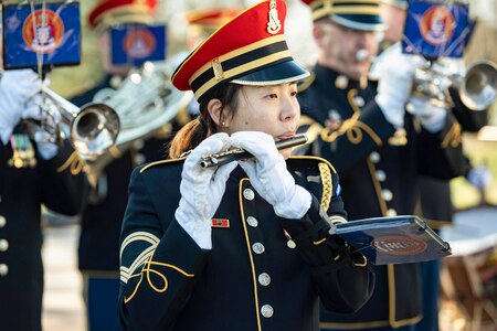 A close-up of a band member in ceremonial uniform playing an instrument as fellow band members perform in the background.