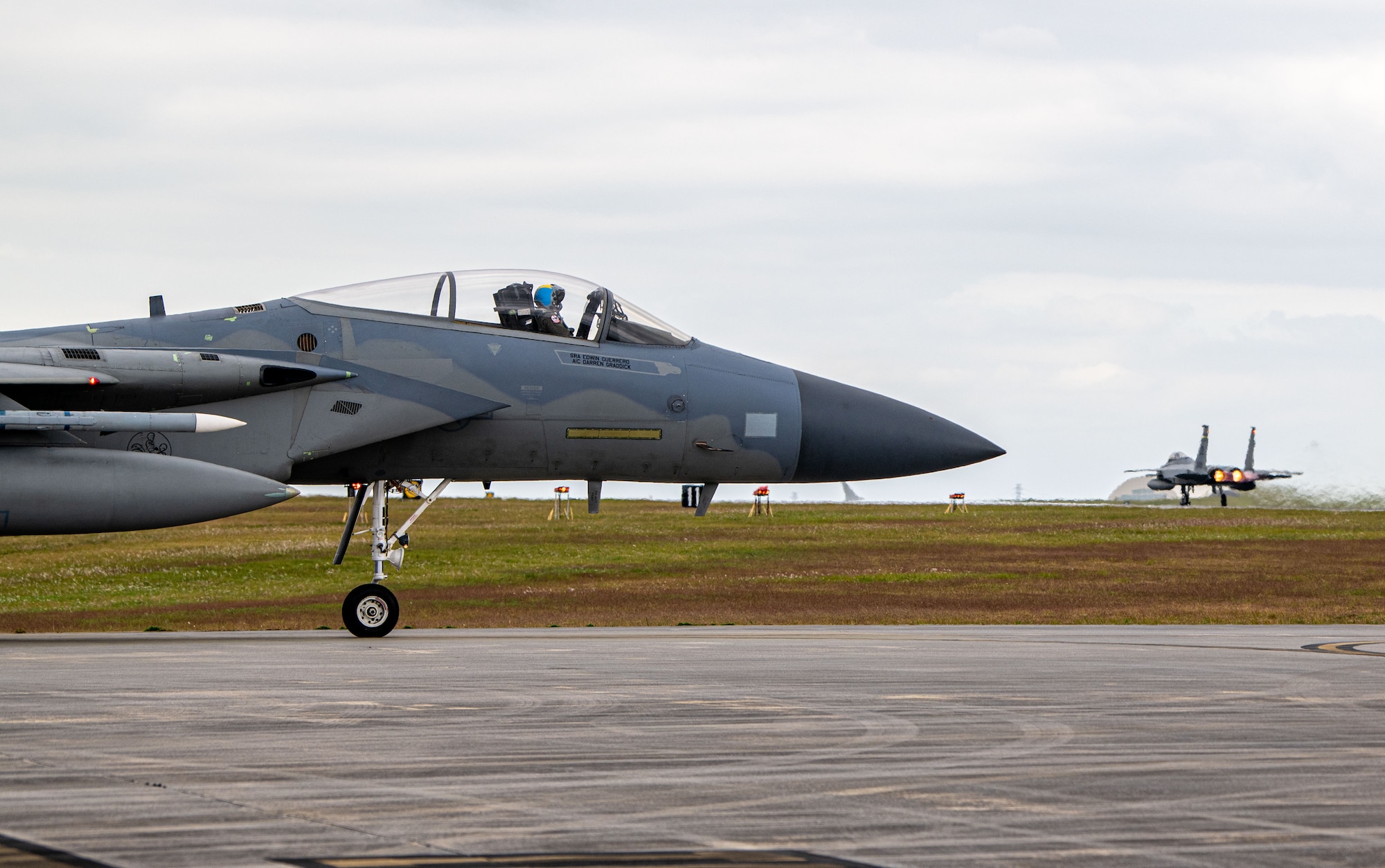 An F-15C Eagle Taxis on a runway while another F-15C Eagle Takes off in the background.