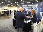 A dark skinned woman and a light skinned man in business attire talk to a light skinned man in business attire at a booth on a showroom floor.