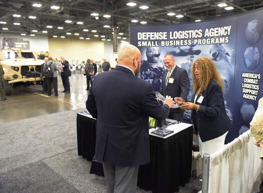 A dark skinned woman and a light skinned man in business attire talk to a light skinned man in business attire at a booth on a showroom floor.