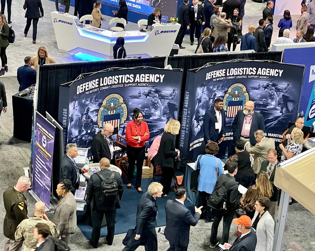 A group of people visit a Defense Logistics Agency booth in a large conference center. Photo shot from above.