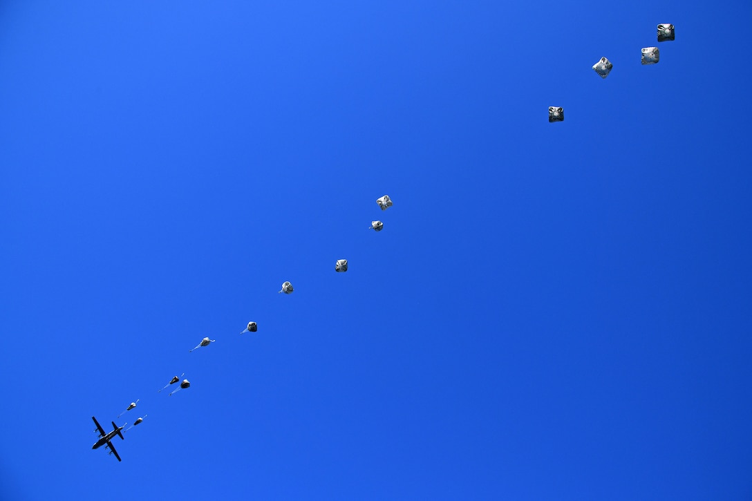 A military aircraft flies as paratroopers descend in parachutes against a bright blue sky.
