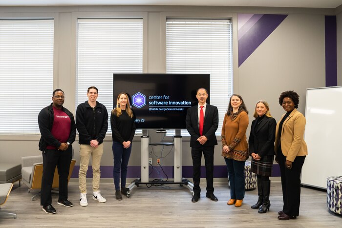 Seven people standing, posing for a photo while in front of a TV.
