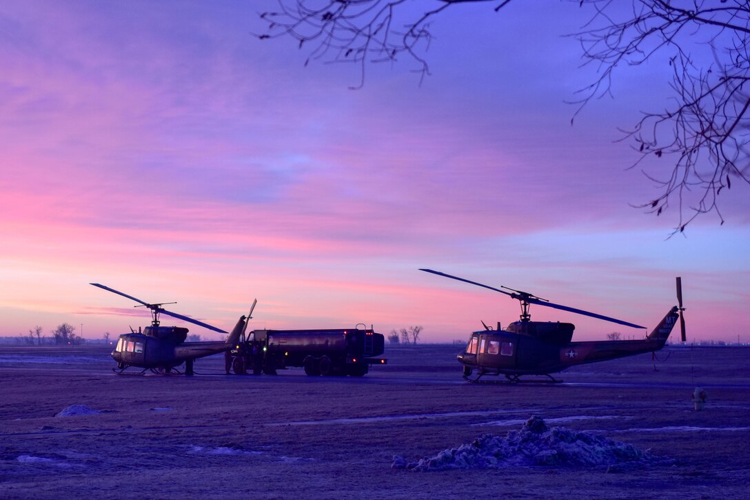 Two helicopters and a large truck sit in a field under a purple sky.