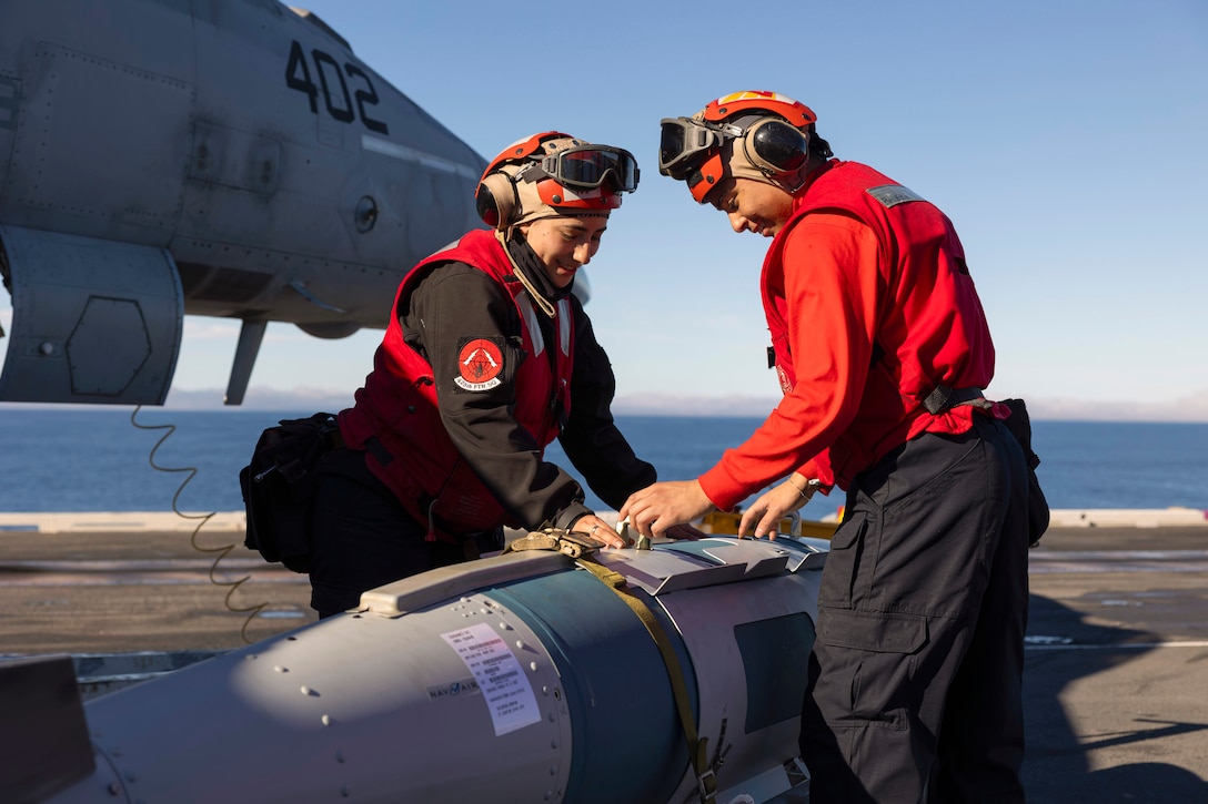 Two sailors work on a weapon connected to an aircraft aboard a ship with a body of water in the background.