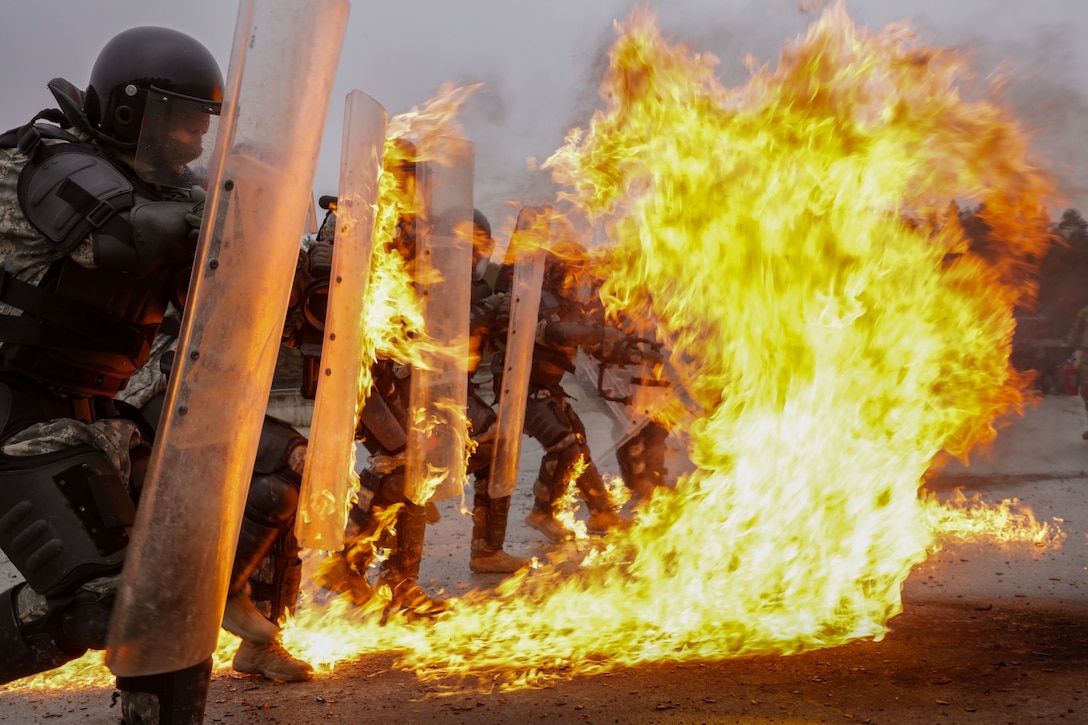Soldiers in police tactical gear hold shields as a fire burns in front of them.