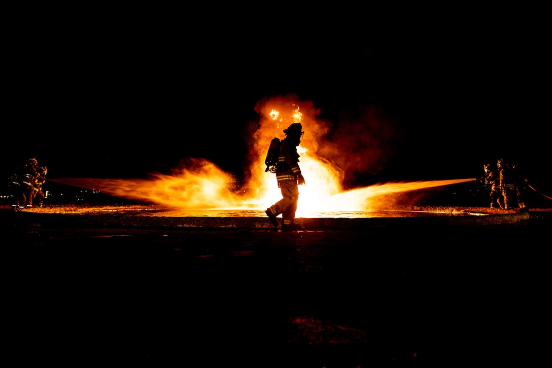 Marines operate fire hoses to extinguish a fire as another Marine walks in front of the fire.