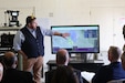 A man stands next to a screen displaying maps and explains the results to three men sitting in chairs and listening.