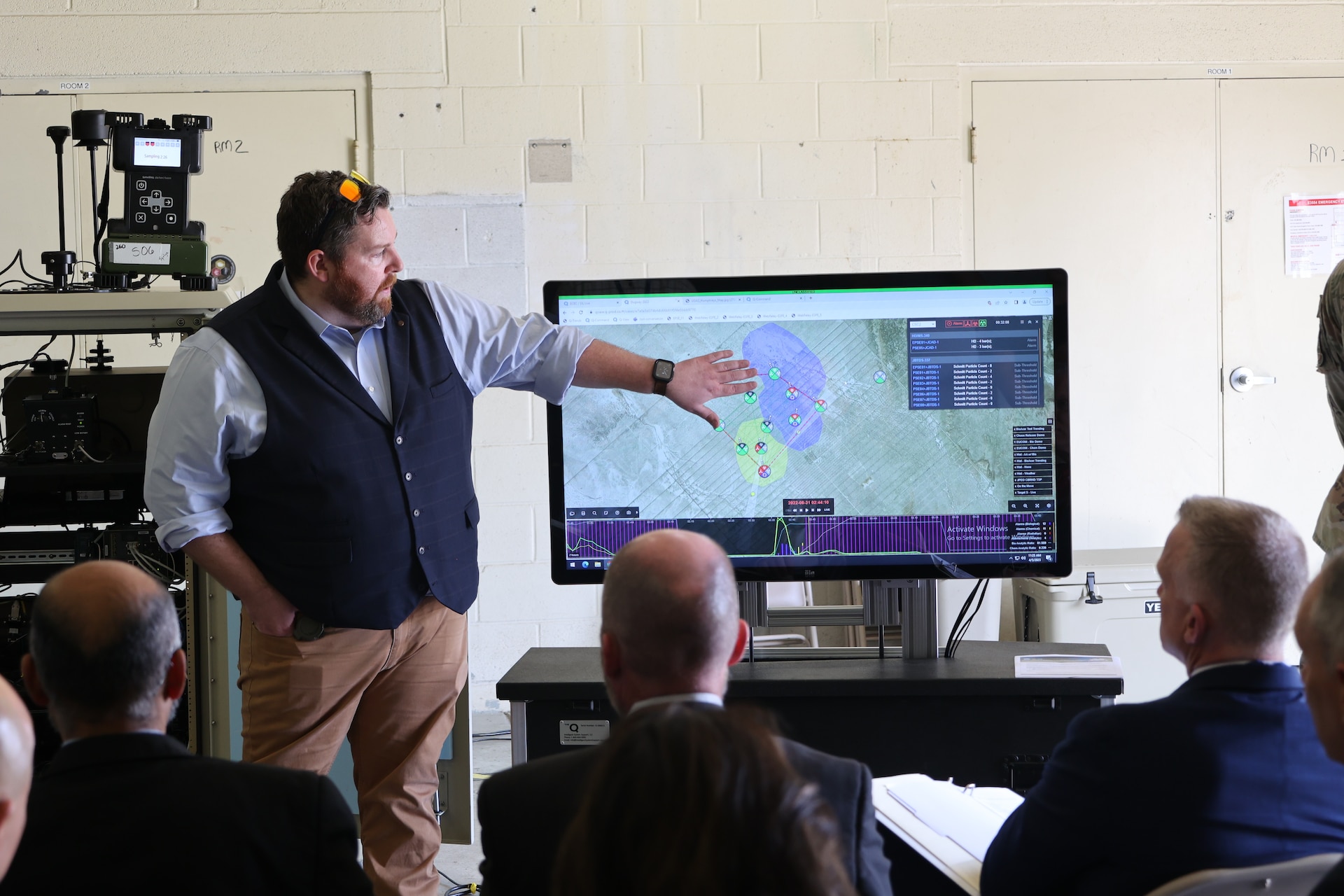 A man stands next to a screen displaying maps and explains the results to three men sitting in chairs and listening.