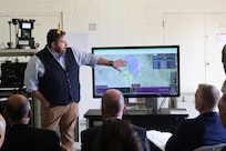 A man stands next to a screen displaying maps and explains the results to three men sitting in chairs and listening.