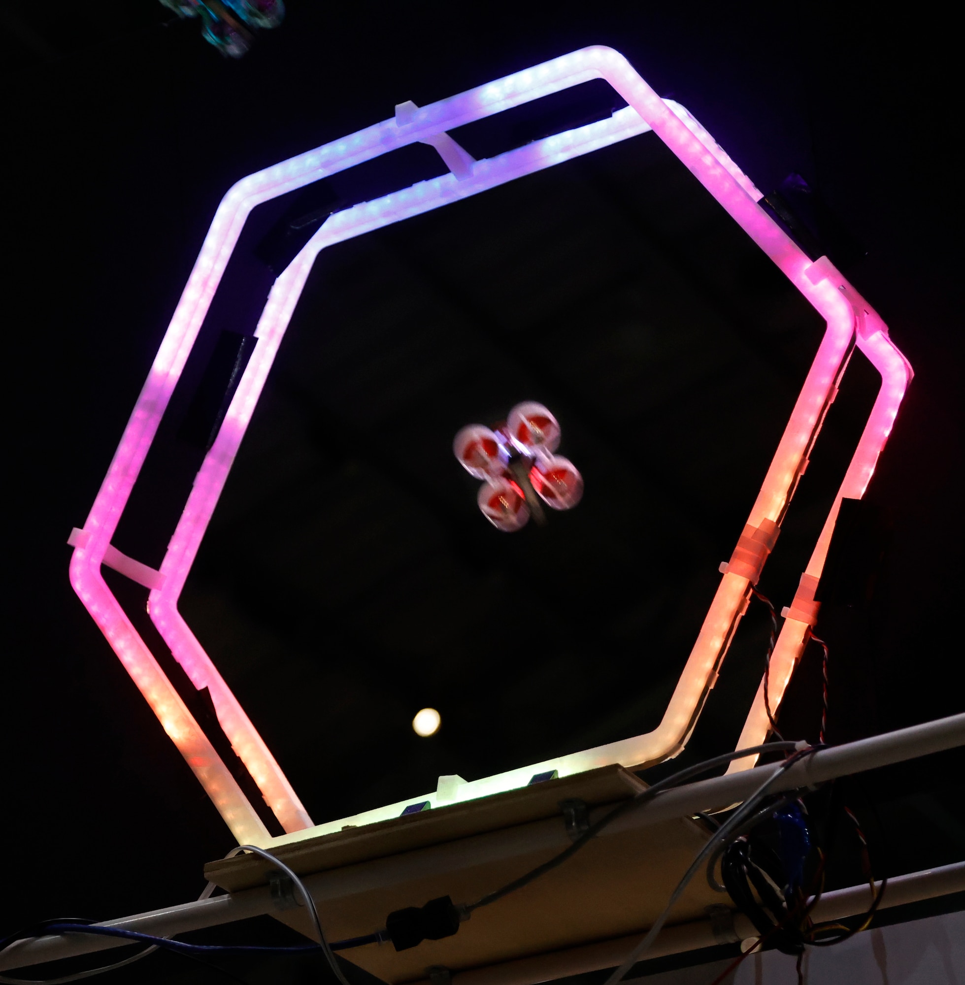 Image of a six-sided opening with a micro drone flying through it. The background is black the the tube is lit up in blue, purple and pink colors.