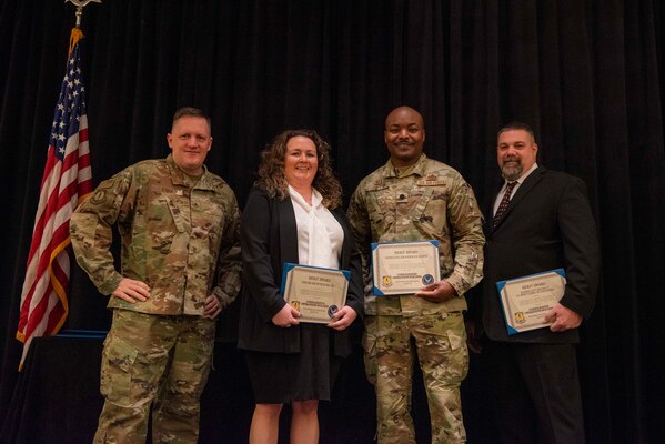 Two men in military uniforms stand with a man and woman, both in black suits and white shirts, holding certificates with an American flag to the left.