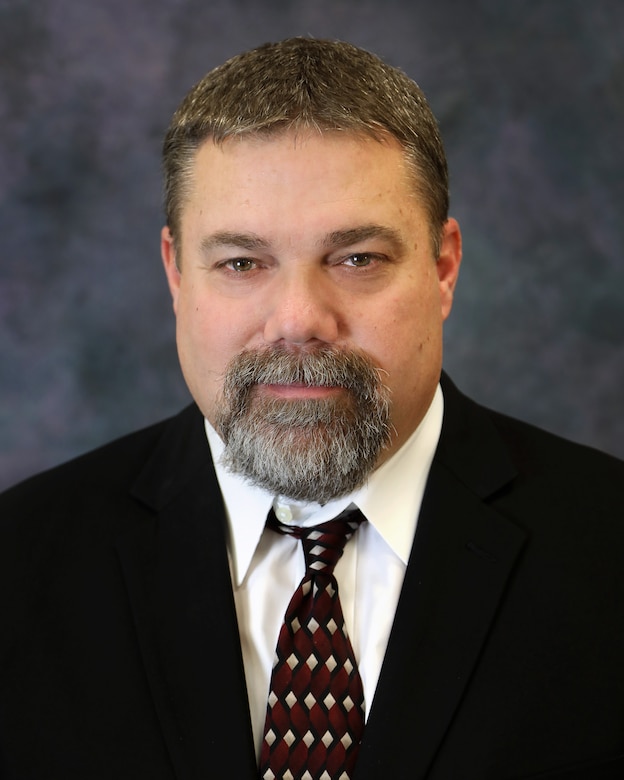 A man with a grey gotee, black jacket, white shirt, and red tie in front of a grey background.