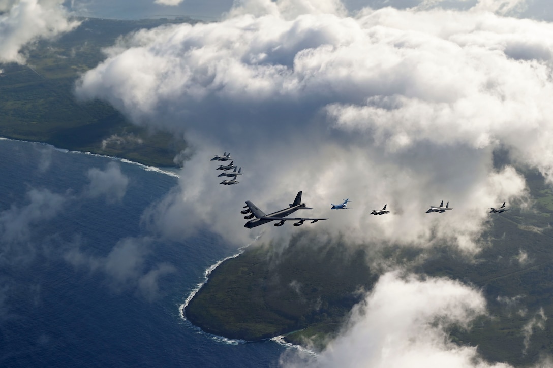 Nine aircraft fly in formation near clouds over an island and body of water.