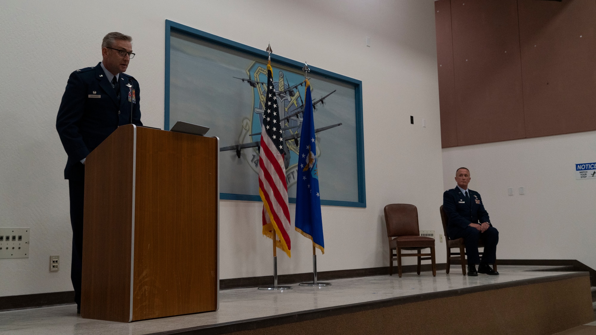 One California Air National Guard officer standing at the podium on a stage while another officer from the same organization is sitting in a chair inside an auditorium on stage.