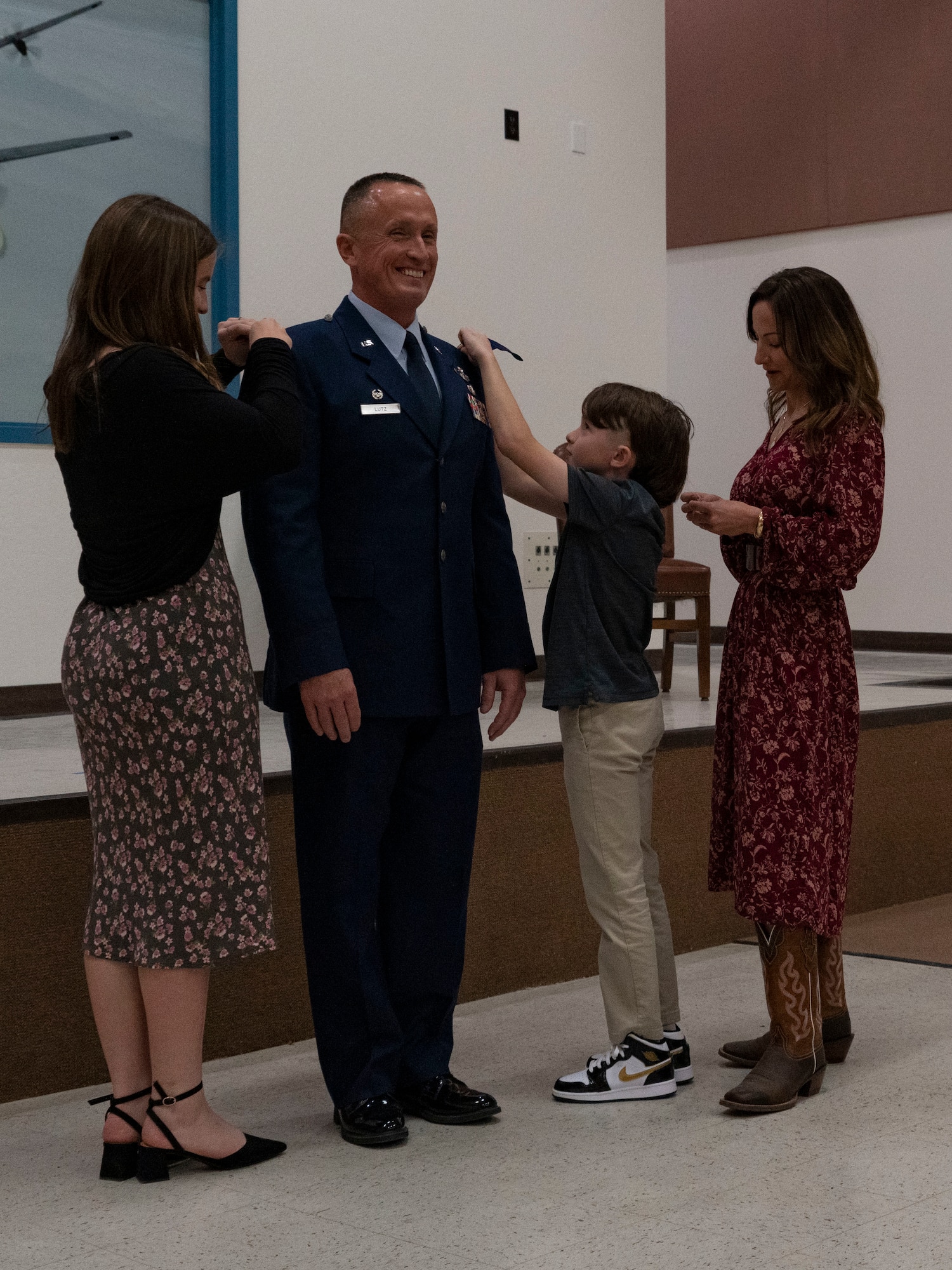 A California Air National Guard officer surrounded by his family that are pinning a colonel rank insignia on his ephialtes that sit at the the top of his shoulders on his Air Force uniform.