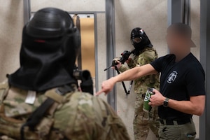 A person in plain clothes giving direction to people in military uniforms wearing protective masks holding rifles.