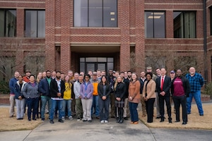 A group of people standing in front of a brick building. They are posed and looking at the camera for a photo.