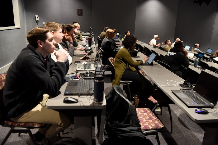 A group of people sitting at long tables with computers in front of them. They are looking forward.