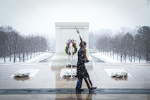 Soldier marching in front of the Tomb of the Unknown Soldier. She is dressed in the Army cold weather uniform and is carrying a rifle on her shoulder. In front of the tomb, there are three wreaths on the ground and another on a stand.