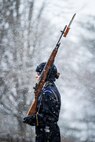 Female Soldier dressed in dark blue winter weather uniform carrying rifle on shoulder with snow falling.