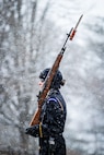 Female Soldier dressed in dark blue winter weather uniform carrying rifle on shoulder with snow falling.