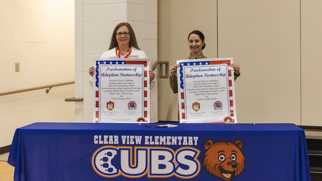 Denise D. Gartner, left, principal of Clear View Elementary School, and U.S. Marine Corps Lt. Col. Laura J. Perazzola-Ash, right, commanding officer, Wounded Warrior Battalion-East (WWBn-E), hold up the proclamation of adoption partnership during an Adopt-a-School ceremony on Feb. 5, 2024 at Clear View Elementary School in Jacksonville, North Carolina.  The Adopt-a-School program gives a military unit the ability to support local schools by assisting with field trips, family nights, Month of the Military Child and other special events. (U.S. Marine Corps photo by Cpl. Jennifer E. Douds)