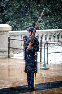 Soldier standing at attention at the Tomb of the Unknown soldier wearing cold weather uniform while carrying rifle in snowy weather conditions.