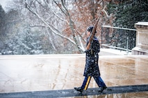 Army Soldier wearing cold weather uniform marching on mat in front of the Tomb of the Unknown Soldier in snowy conditions with a rifle in hand.