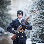 U.S. Army Soldier in winter uniform with sunglasses holding rifle while marching in snowy conditions.