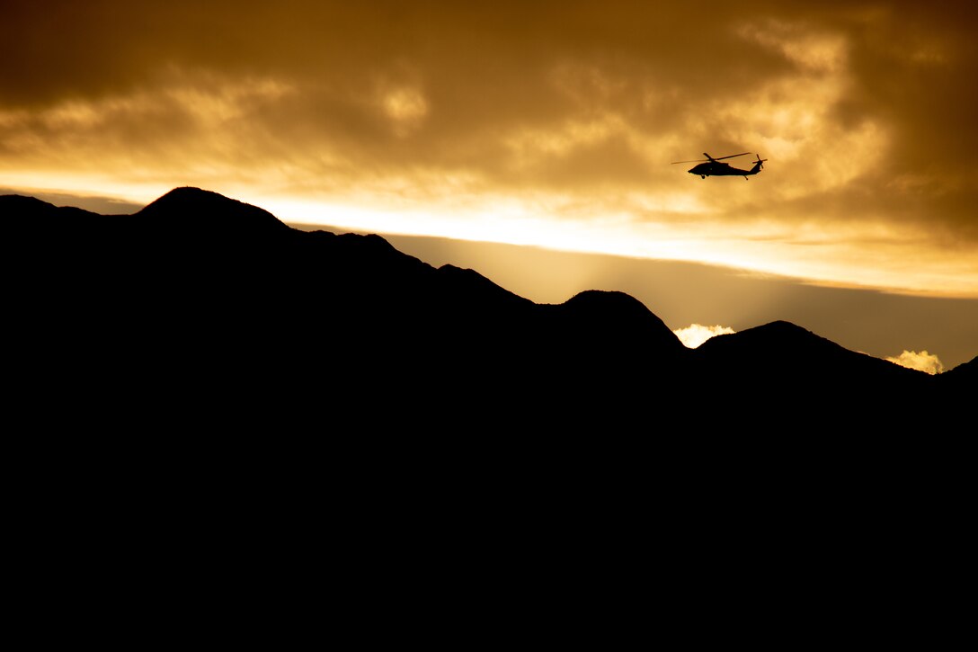 A helicopter is silhouetted against a cloudy yellow sky flying above darkened mountains.