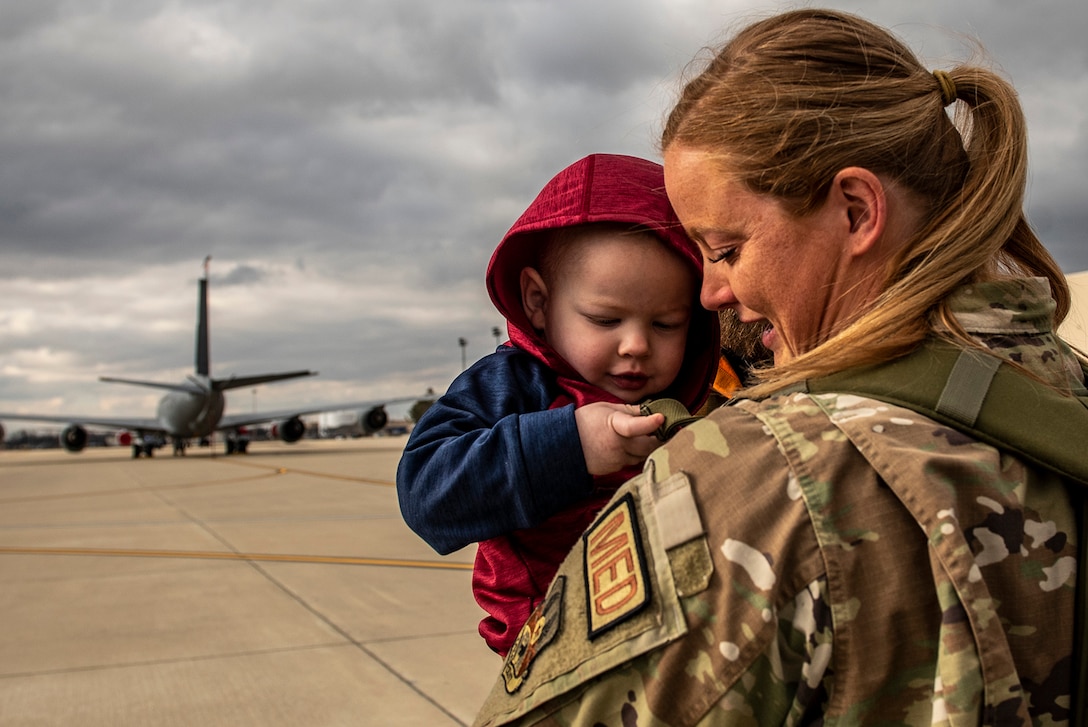 A uniformed service member holds a child. A parked aircraft can be seen in the background.