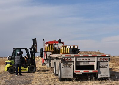 a green forklift loads a pallet of brass colored shells onto a flatbed semi truck. This operation is taking place outside on gravel and grass covered  ground