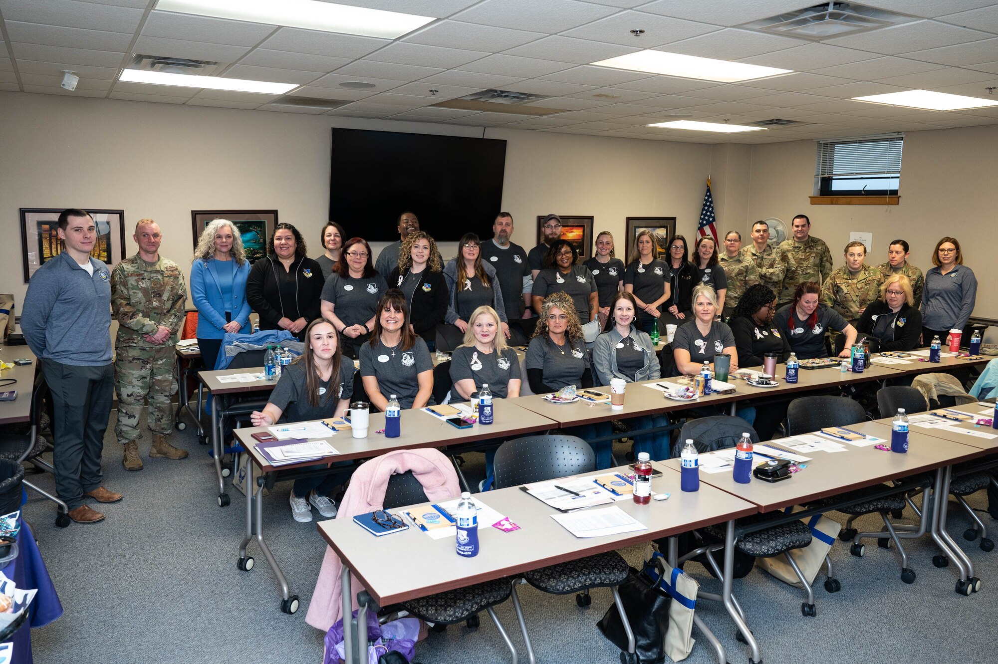A group photo of men and women in rows, some sitting and some standing in a classroom-like setting. The majority are wearing dark gray and black polos.