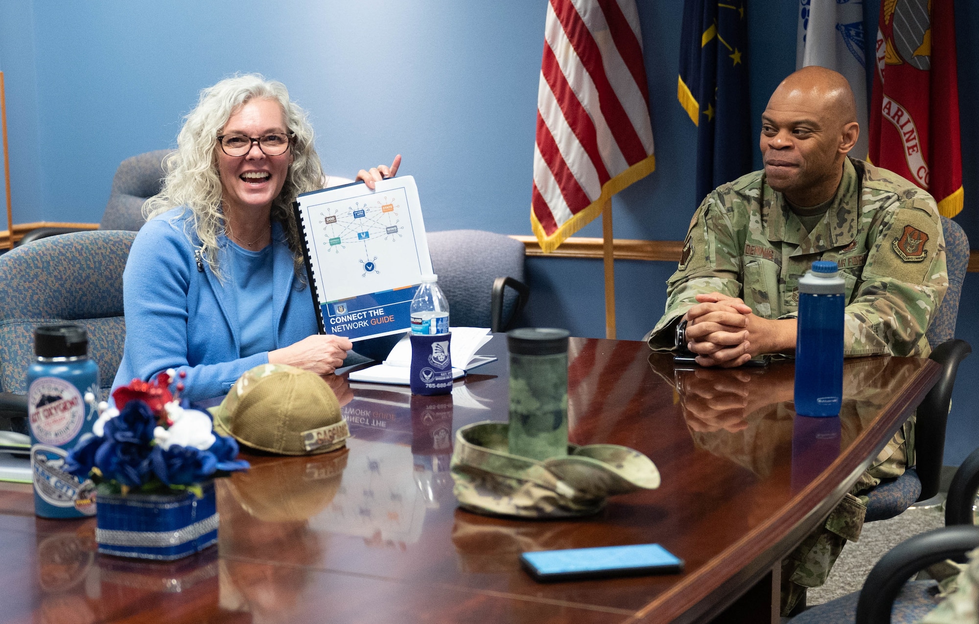 A woman holds up a spiral-bound book that reads "Connect the Network Guide" while sitting down at a table. At right, a man in a military uniform smiles at her as she presents.